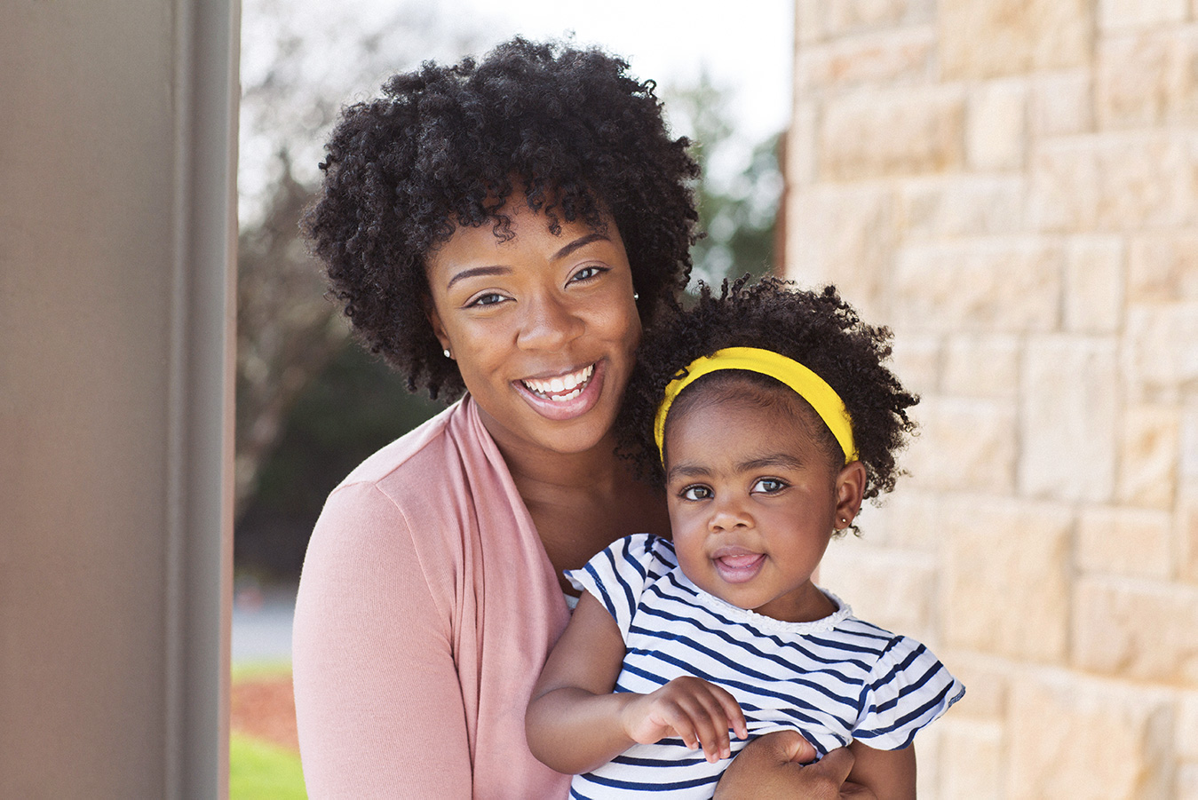 A young Black mother and her toddler daughter smiling at the camera, sitting in front of their home